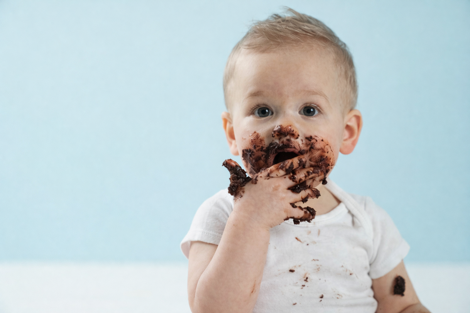Bébé d’environ un an avec le visage et les mains couverts de mousse au chocolat, dégustant un dessert fait maison, en format paysage sur fond bleu clair.