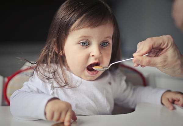 Bébé assise dans une chaise haute, recevant une cuillère de purée par un adulte pendant un repas de diversification alimentaire.