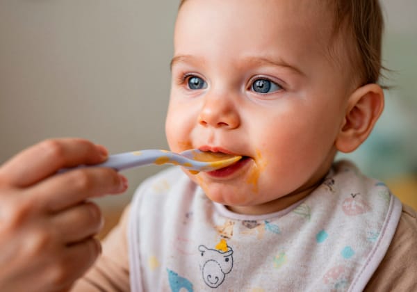Bébé aux yeux bleus mangeant une purée à la cuillère, portant un bavoir coloré