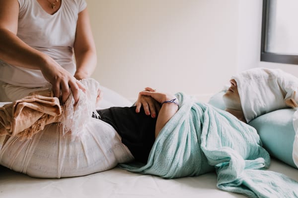 Femme allongée recevant un soin Rebozo, enveloppée dans des châles colorés, les yeux couverts d’une serviette, dans une atmosphère apaisante et chaleureuse.