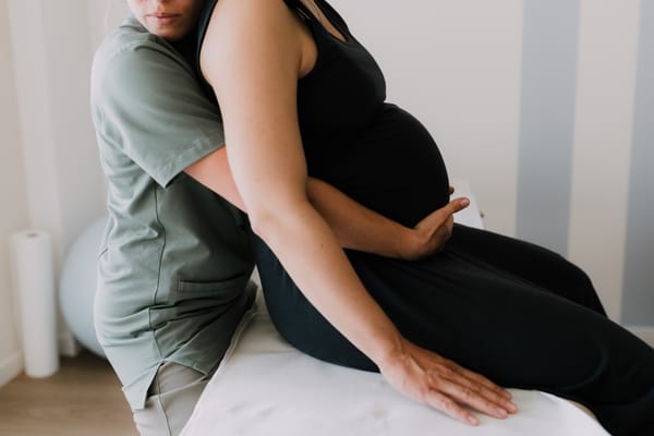 Femme enceinte recevant un soin ostéopathique ou massage prénatal, assise sur une table d’examen, soutenue par une professionnelle de santé pendant la grossesse.
