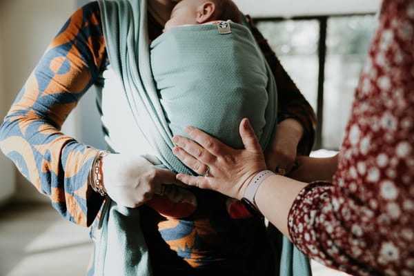 Bébé porté en écharpe de portage physiologique contre son parent, accompagné par une professionnelle, illustrant une position sécurisée et respectueuse du développement du nourrisson.